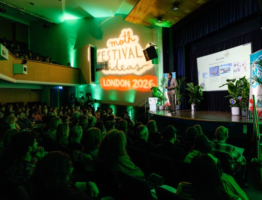 César Rodríguez-Garavito presents onstage to a full audience during the MOTH Festival of Ideas London 2026. Green lighting fills the auditorium while a large illuminated festival projection appears on the wall beside the stage. Presentation slides are displayed on a screen surrounded by plants and festival banners.